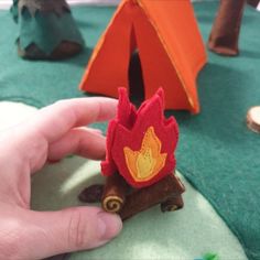 a hand is holding a small campfire brooch in front of a tent on the ground