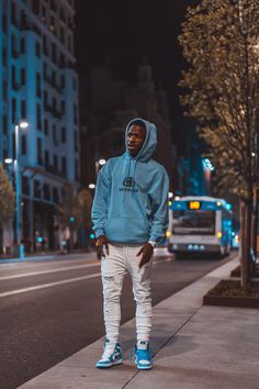 a man in a blue hoodie and white pants stands on the sidewalk at night