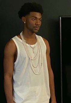 a man in white shirt and pearls standing next to a black wall with his hands in his pockets