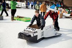 two young men riding snow boards on top of snow covered ground with people in the background