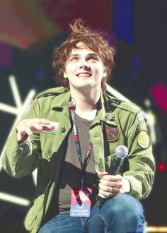 a young man holding a microphone in his right hand and wearing a green jacket with patches on it