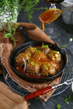 meatballs with sauce and onions in a black bowl on a plate next to a red handled utensil