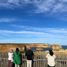 three people looking out over the ocean from a wooden fence with cliffs in the background