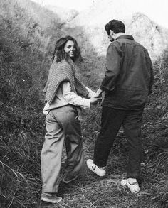 two people holding hands while standing in the grass near some bushes and trees, with mountains in the background