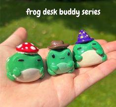 two green frog figurines with hats on their heads sitting in the palm of someone's hand