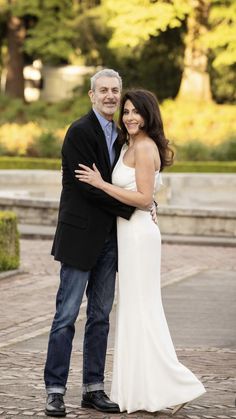 an older man and woman posing for a photo in front of some trees with their arms around each other