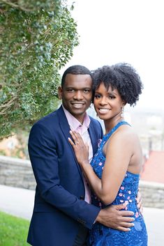 a man and woman standing next to each other in front of a tree with their arms around each other