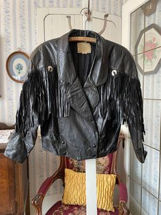 This fantastic coat from The Boutique of Leathers is made from genuine leather in classic black. It has two outside pockets and fastens at the waist with four snaps, with black leather fringe on the front and back yokes and shoulders, with round silver accents. The measurements, taken with the jacket lying flat, are: shoulder to shoulder, 17 inches; armpit to armpit, 20 inches; sleeves, 22 inches; length, 25 inches in front and 21 1/2 inches in back; bottom edge, 18 inches. In very good condition. Black Leather Outerwear With Fringe, Black Leather Fringe Outerwear, Black Leather Jacket With Fringe For Winter, Black Fringed Leather Jacket For Fall, Vintage Winter Leather Jacket With Fringe, Vintage Leather Jacket With Fringe For Winter, Vintage Fringe Leather Jacket For Winter, Fall Biker Leather Jacket With Fringe, Edgy Leather Jacket With Fringe For Winter