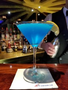 a blue cocktail in a martini glass with a lemon slice on the rim and a bartender behind it