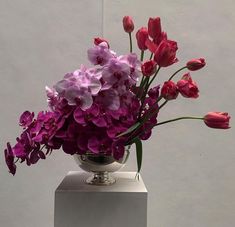 a silver vase filled with purple and red flowers on top of a white pedestal next to a wall