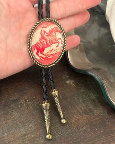 This awesome bolo tie has a vintage cowboy print with a clear resin coating and metal backing. Handmade in our shop! The cord is black and genuine leather. Our bolos pair nicely with many of our belt buckles! They make wonderful gifts.    The western bolo tie rope length is 39' ;  pendant size is 1 1/2'' x 1 1/4'' Vintage Black Bolo Ties For Gift, Vintage Black Bolo Ties As Gift, Vintage Black Jewelry For Western-themed Events, Vintage Bolo Ties With Adjustable Length For Rodeo, Vintage Bolo Tie With Adjustable Length For Western Events, Vintage Bolo Ties With Adjustable Length For Western-themed Events, Vintage Style Adjustable Bolo Ties As Gifts, Brown Bolo Ties As A Gift, Brown Bolo Tie For Gift