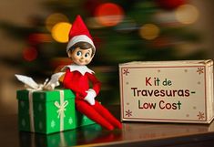 an elf sitting on top of a green box next to a christmas tree and a sign that says kit de travesuras - low cost