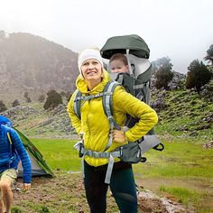 two people with backpacks standing next to a tent in the mountains, one carrying a baby