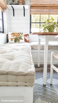 a white bed sitting under a window next to a wooden table and two stools