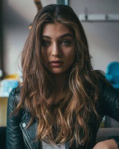 a woman with long hair sitting on a couch in front of a window wearing a black leather jacket