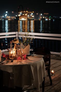 the table is set on the deck by the water at night with candles lit up