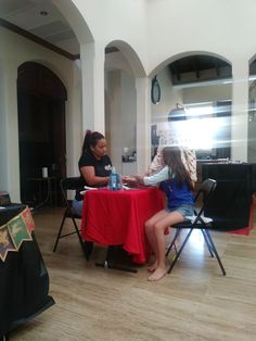 two women sitting at a table talking to each other in a large room with wooden floors