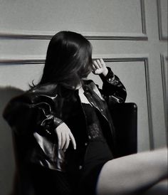 a woman sitting in a chair talking on a cell phone while wearing a leather jacket