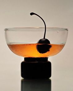 a glass bowl filled with liquid and a black cherry on the top, sitting on a reflective surface