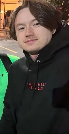 a man with long hair wearing a black hoodie and smiling at the camera while sitting on a bench