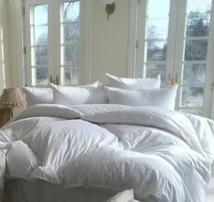 a bed with white comforters and pillows in a room next to two large windows