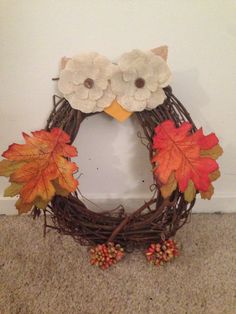 an owl wreath with leaves and flowers on the front is sitting next to a wall