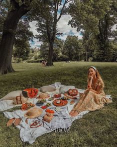 a woman sitting on a blanket in the middle of a park with food spread out