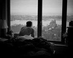 a man sitting on top of a bed in front of a window looking out at the city