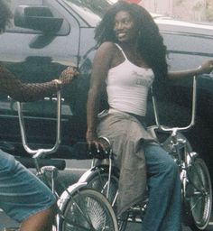 two women sitting on bicycles in front of a car and another woman standing next to them