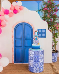 a blue and white table topped with a cake next to a wall covered in balloons