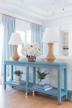 a blue console table with two lamps and flowers on it in front of a window