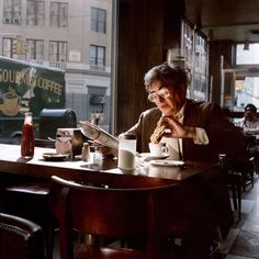 a man sitting at a table in front of a window reading a newspaper and drinking coffee