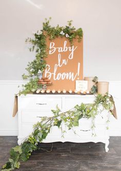 a baby in bloom sign on top of a dresser with greenery and potted plants