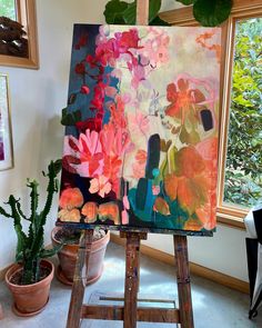 an easel with flowers painted on it in front of a window and potted plants