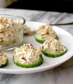 small cucumbers with tuna salad on them sit on a white plate next to a glass bowl