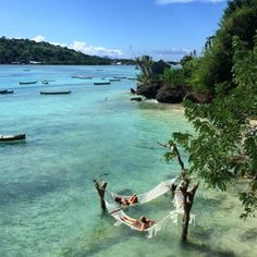 two people laying in a hammock on the beach with boats in the water