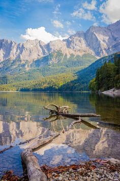 a lake surrounded by mountains and trees with water in the foreground on a sunny day