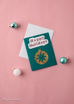 a green and gold holiday card on top of a pink surface next to christmas ornaments