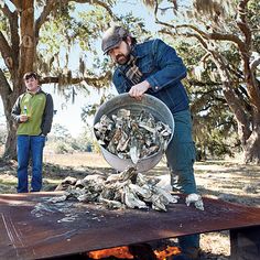 two men standing next to each other in front of a fire pit filled with oysters