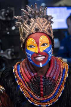 a woman with painted face and colorful jewelry around her neck, standing in front of a television screen