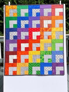 a multicolored quilt is hanging on a white stand with trees in the background