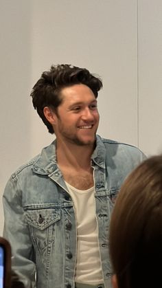 a man in a jean jacket is smiling and talking to the camera while people are looking at him