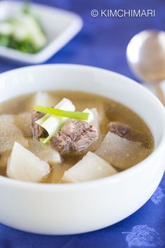 a white bowl filled with soup and meat on top of a blue table cloth next to silver spoons