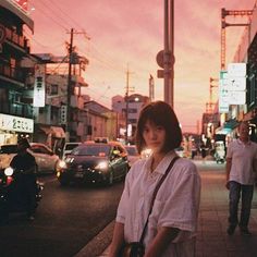 a woman standing on the side of a street next to cars and people walking by