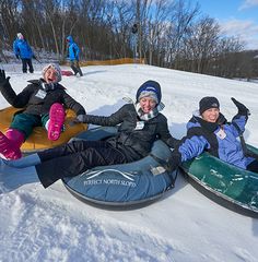 three people sitting on snow sleds in the snow with one person laying down