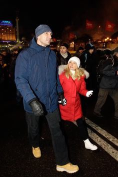 a man and woman standing in the middle of a street at night wearing winter clothes