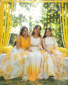 three women in yellow and white dresses sitting on a bench with flowers hanging from the ceiling