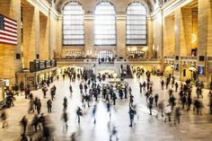 many people are walking around in the lobby of a train station with large windows and an american flag hanging from the ceiling