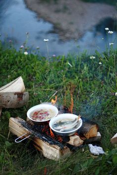 a campfire with plates and bowls on it sitting in the grass next to a river