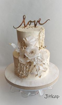 a three tiered cake with white flowers and gold lettering on top that says love